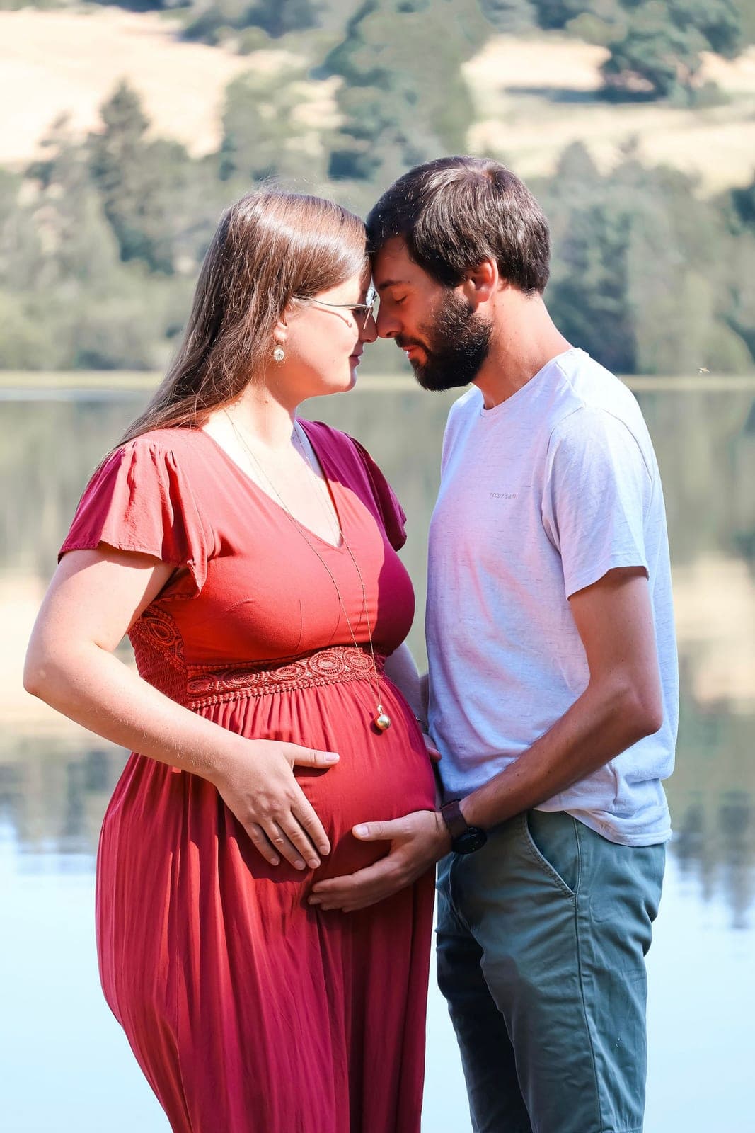 Une future maman en robe framboise, la main posée sur son ventre, au bord d’un lac.