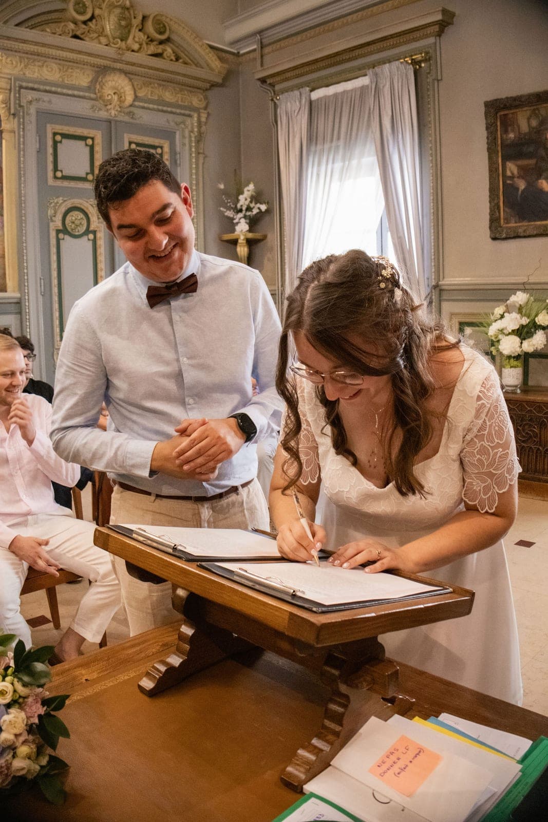 La mariée signe le registre à la mairie, son mari penché près d’elle.