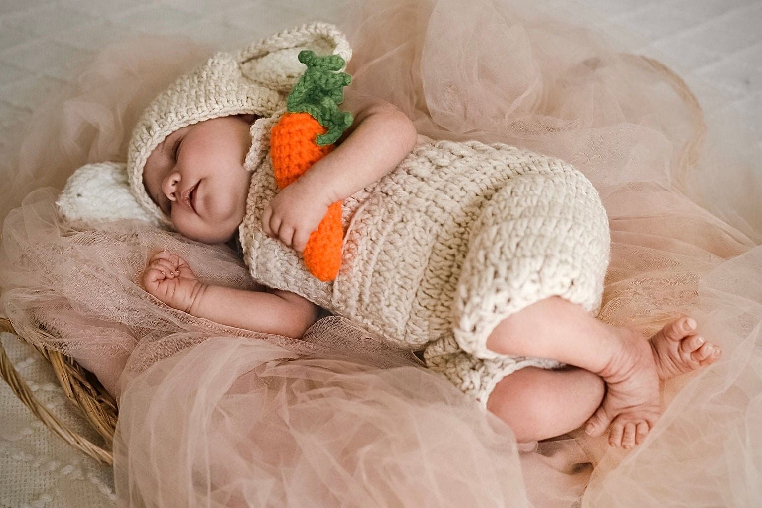 Nouveau-né endormi en bonnet et salopette de laine écrue, une carotte au crochet dans les bras, sur un voile de tulle rose.