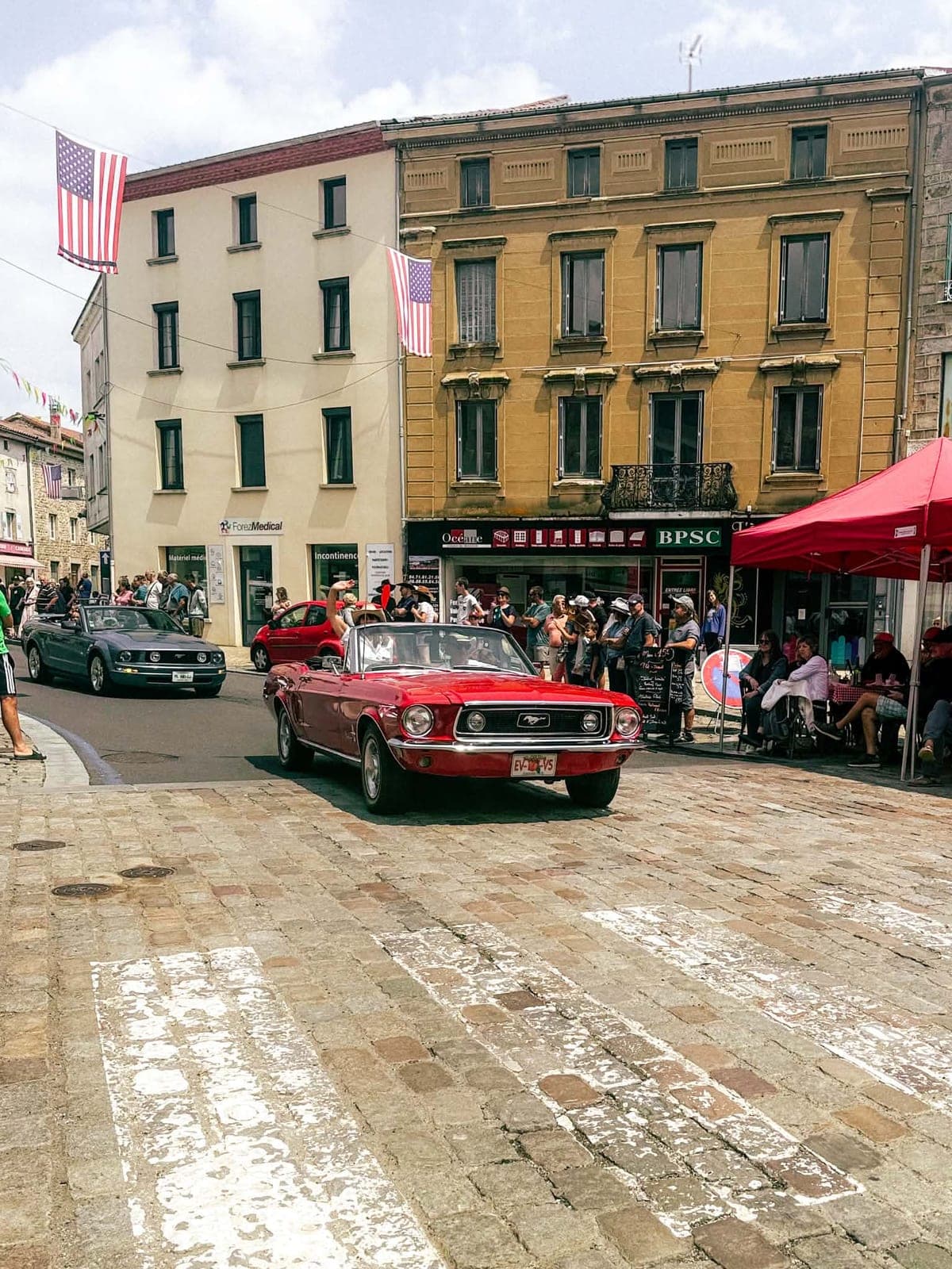 Une Ford Mustang rouge décapotable remonte la place pavée du village, drapeaux américains suspendus au-dessus de la rue.