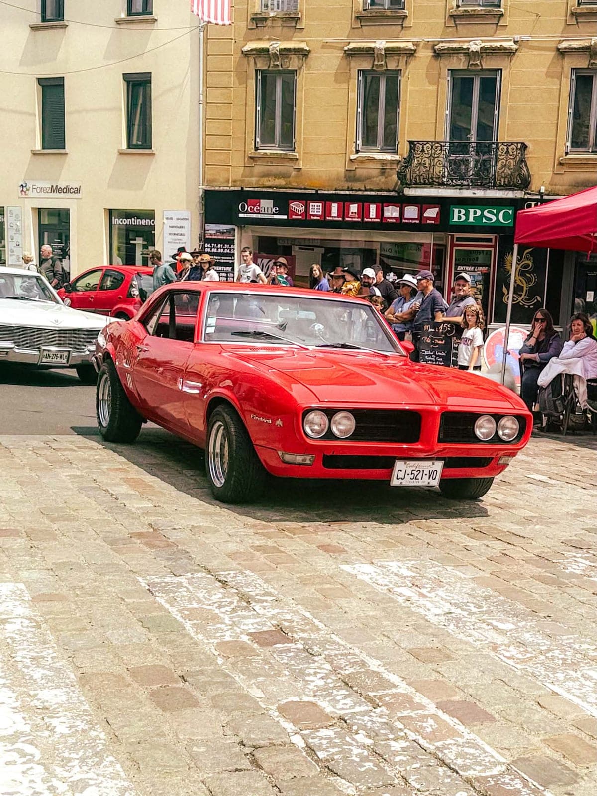 Une Pontiac Firebird rouge exposée sur les pavés, devant les commerces du centre-bourg.