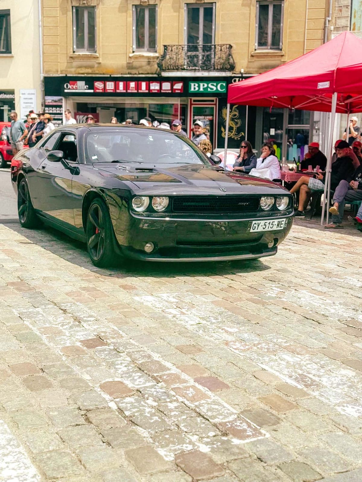 Une Dodge Challenger noire au milieu de la place, les terrasses installées juste derrière.
