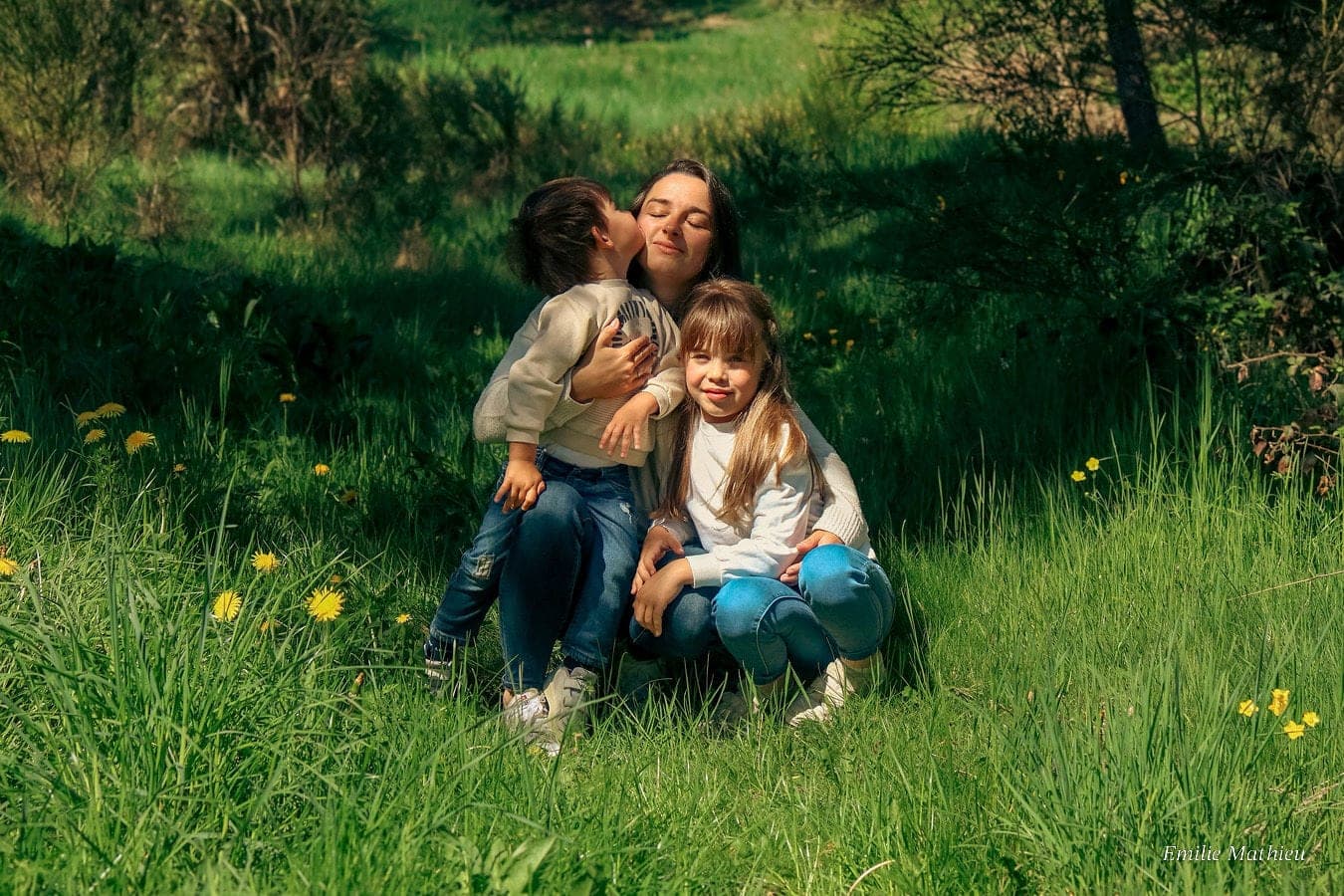 Une maman et ses deux enfants assis dans l’herbe fleurie, un baiser sur la joue.