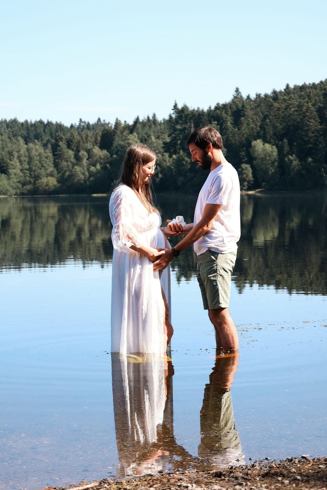 Les pieds dans l’eau du lac, un couple se tient les mains autour du ventre rond.