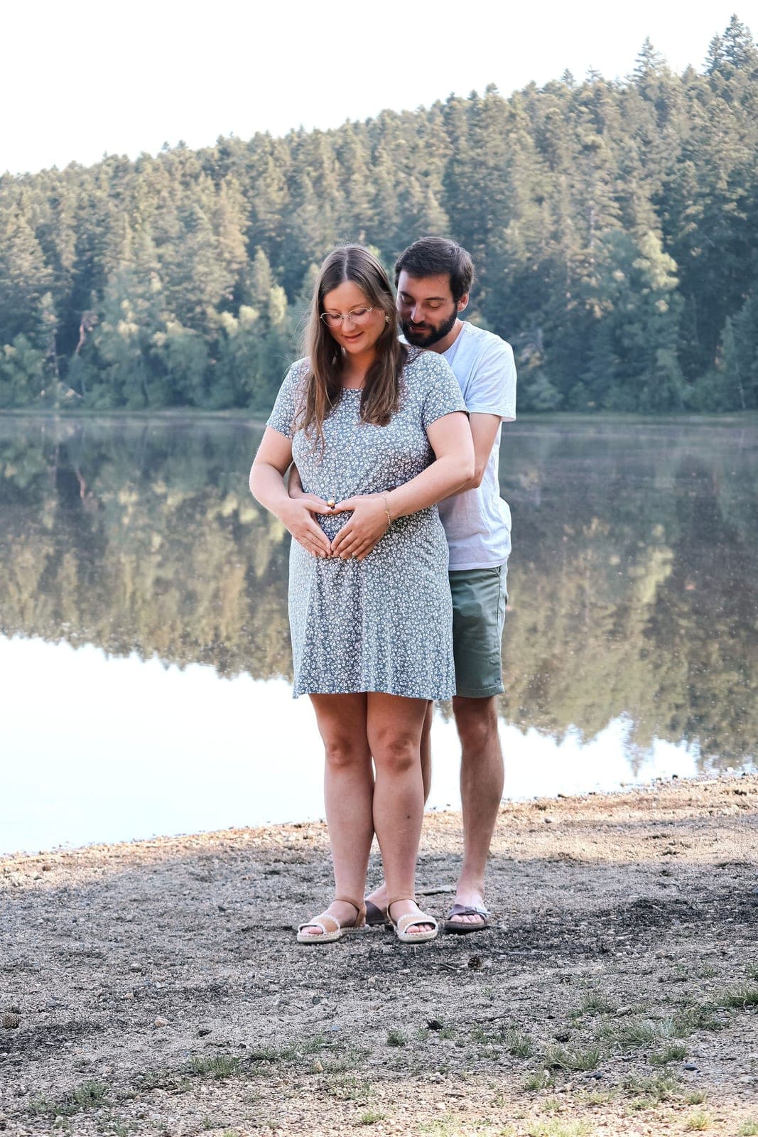 Un couple attend son bébé au bord du lac, mains réunies sur le ventre.