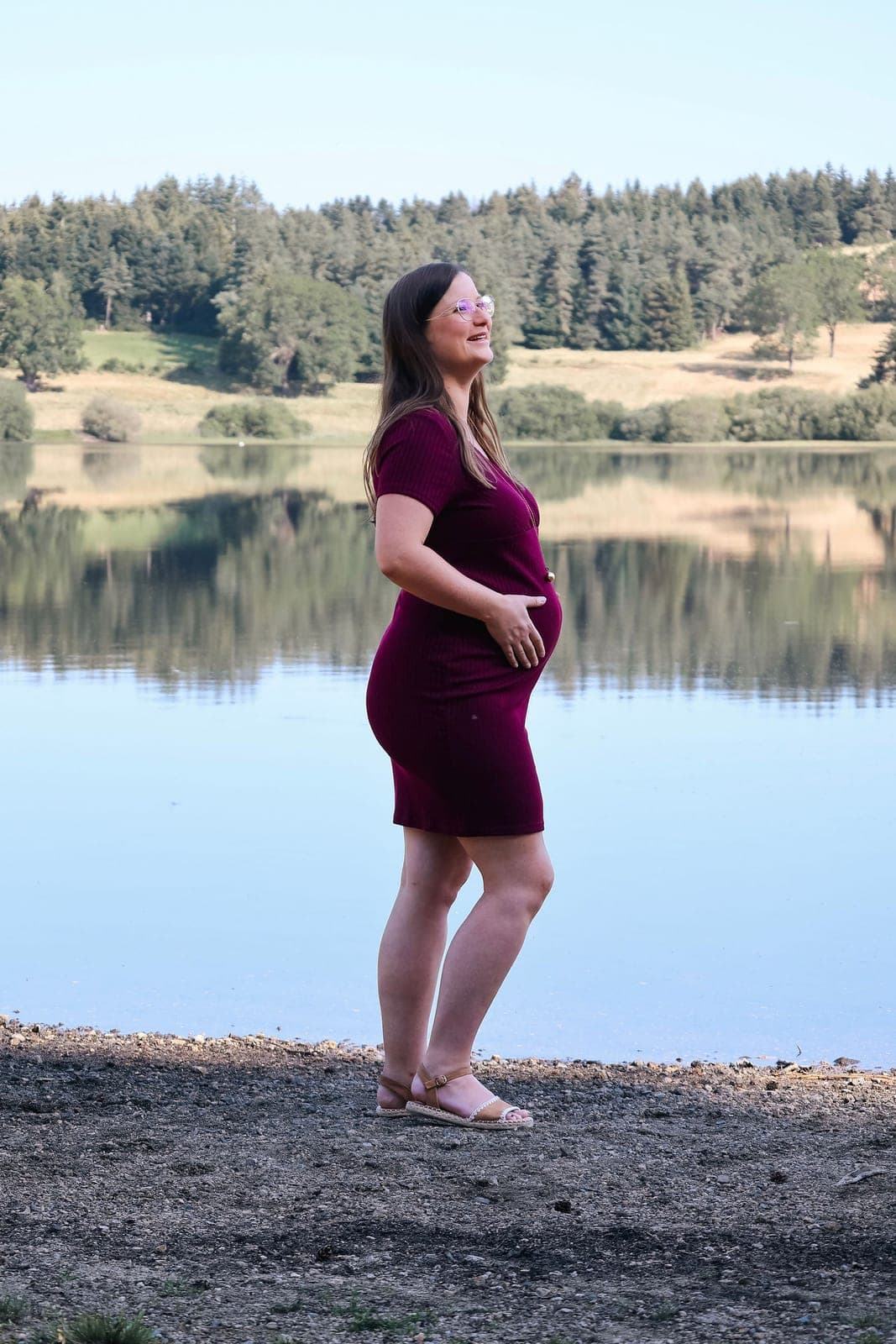 Future maman de profil au bord du lac, en robe framboise.