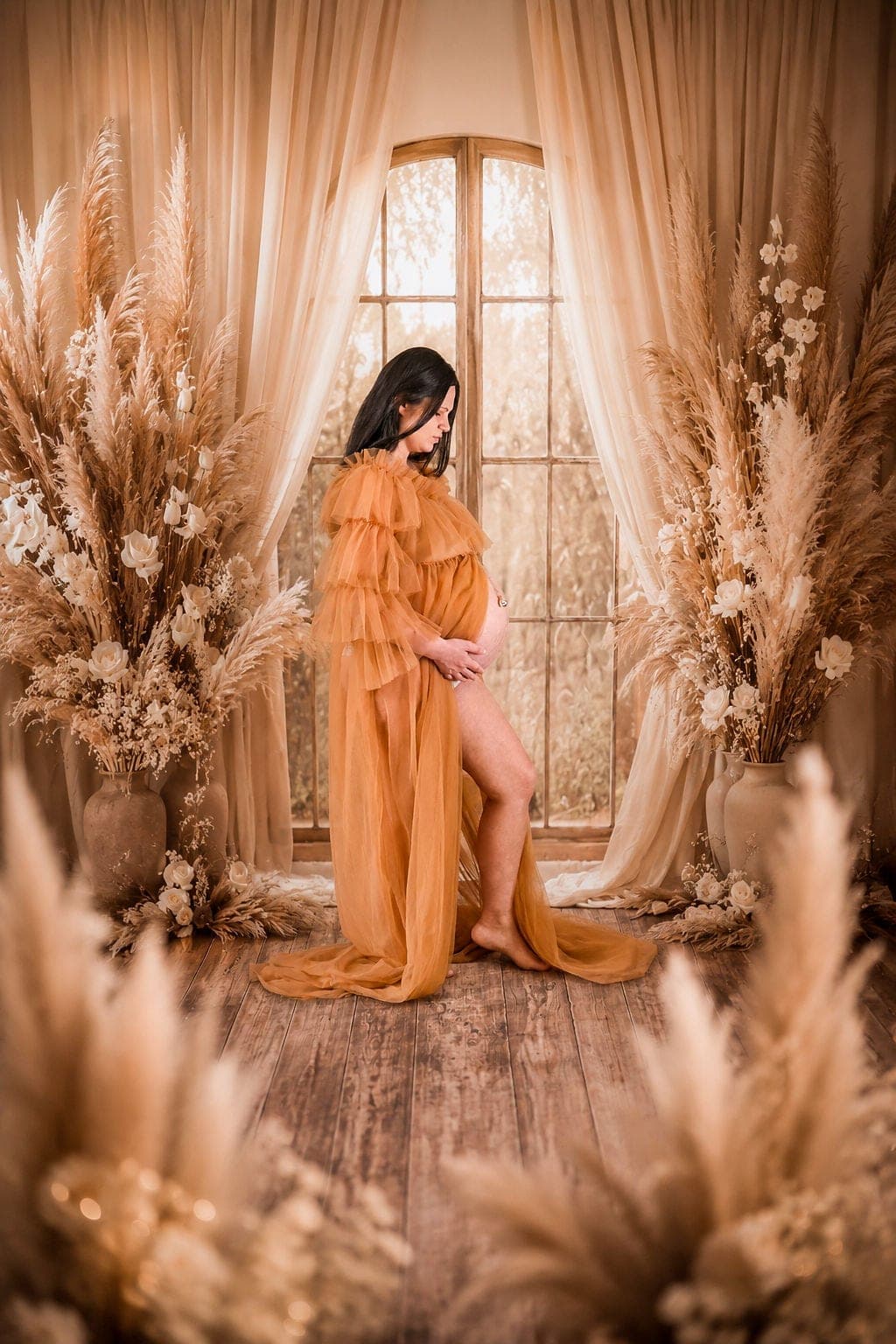 Future maman en robe safran devant la fenêtre du studio, longue traîne au sol.