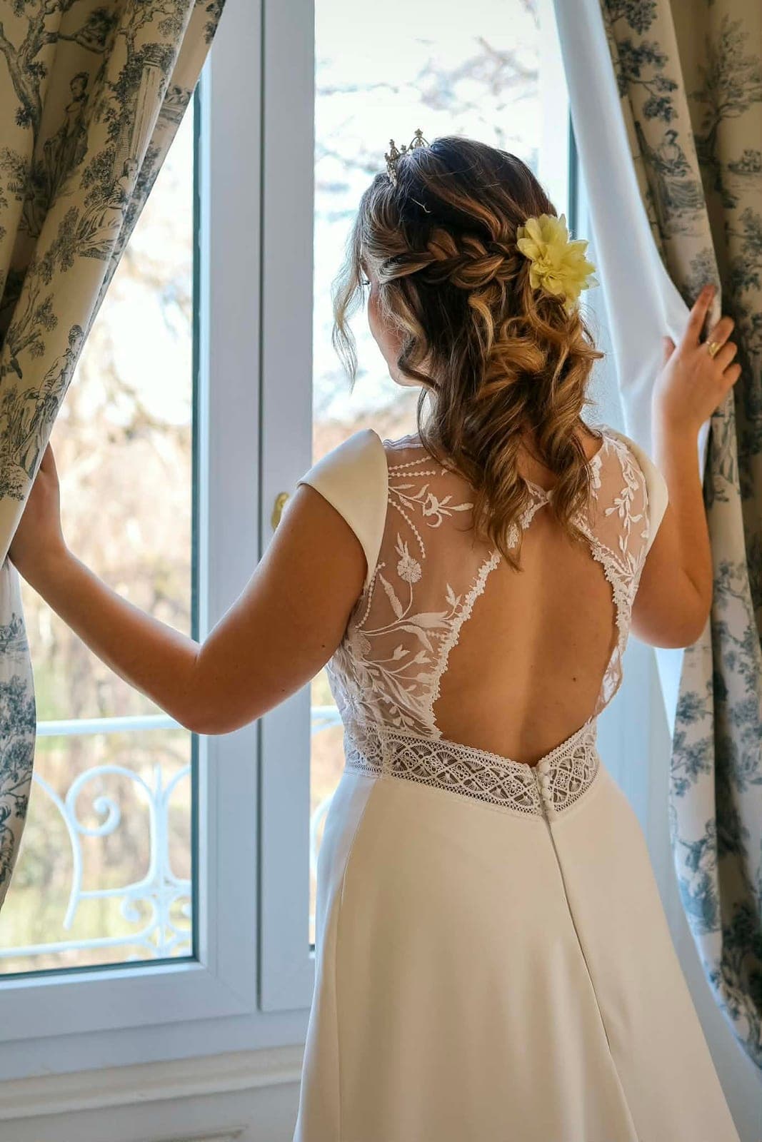 La mariée de dos devant la fenêtre, dos en dentelle et fleur dans les cheveux.